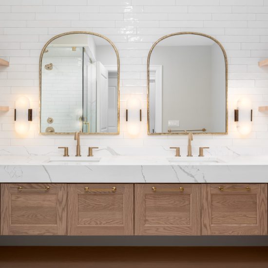 A bathroom detail with gold faucets, light fixtures, and mirrors, a wood cabinet, large marble slab countertop, and a subway tile wall.